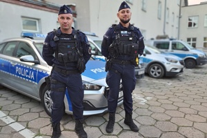 Dwaj policjanci stoją na dziedzińcu komendy na tle radiowozów